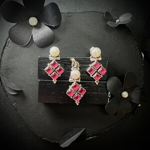 May include: A set of silver earrings and a pendant featuring a square design with four pink gemstones and a pearl accent.
