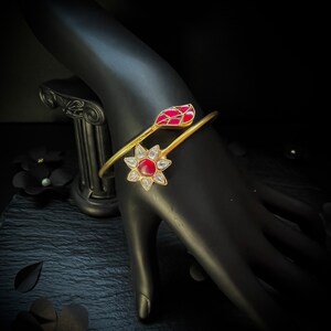Könnte beinhalten: Ein goldenes Armreif-Armband mit einem roten und weißen Blumendesign. Das Armband ist offen und hat auf einer Seite ein Blattdesign.