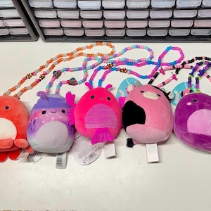 Peut inclure: Cinq colliers avec des jouets en peluche de différentes couleurs. Les jouets comprennent une pieuvre rouge, une créature violette et bleue, un insecte rose, une vache rose avec une tache noire et une créature violette. Chaque jouet est attaché à un collier de perles.