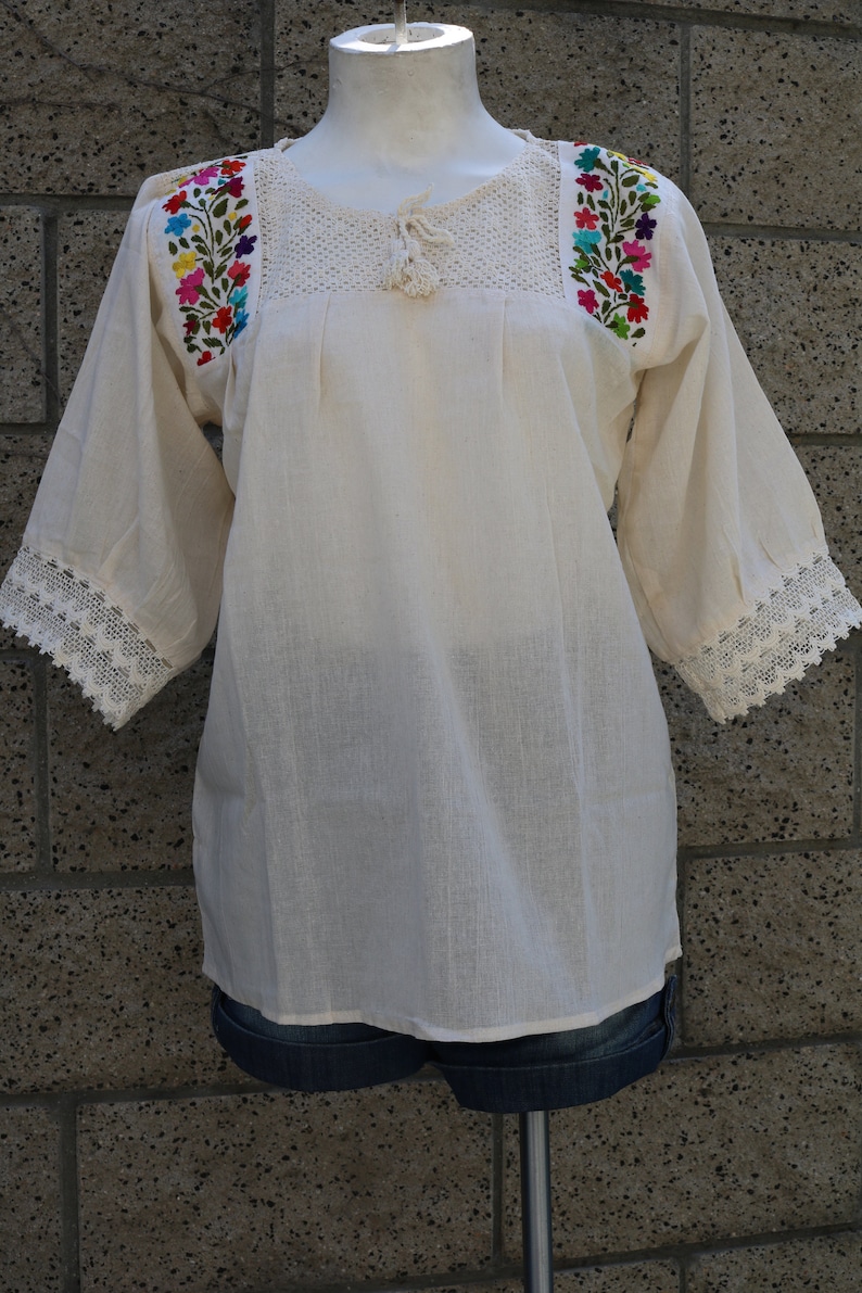 May include: A white cotton blouse with embroidered floral details on the shoulders and lace trim on the sleeves.