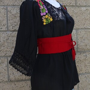 May include: Black embroidered blouse with a red belt and a lace trim. The embroidery features colourful flowers.