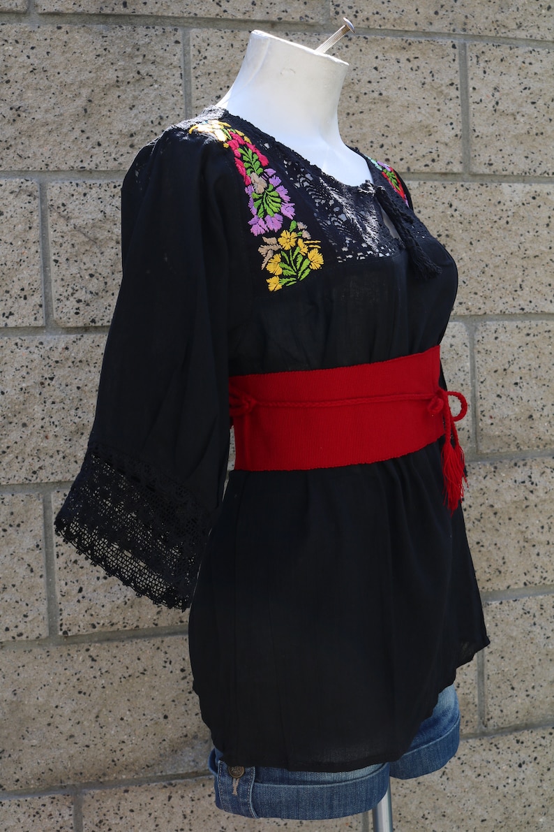 May include: Black embroidered blouse with a red belt and a lace trim. The embroidery features colourful flowers.