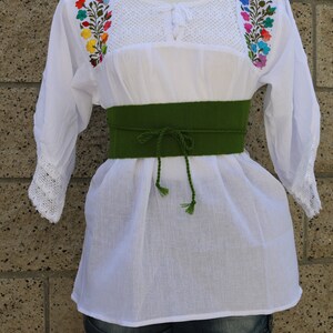 May include: White embroidered blouse with colourful floral designs on the shoulders and a green fabric belt.