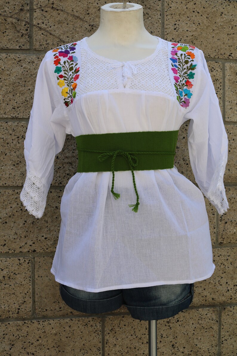 May include: White embroidered blouse with colourful floral designs on the shoulders and a green fabric belt.