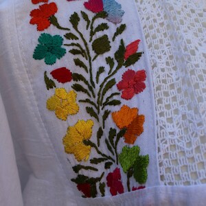 May include: White cotton fabric with colourful embroidered flowers and green leaves. The fabric has a delicate crochet trim.