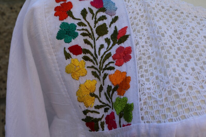 May include: White cotton fabric with colourful embroidered flowers and green leaves. The fabric has a delicate crochet trim.