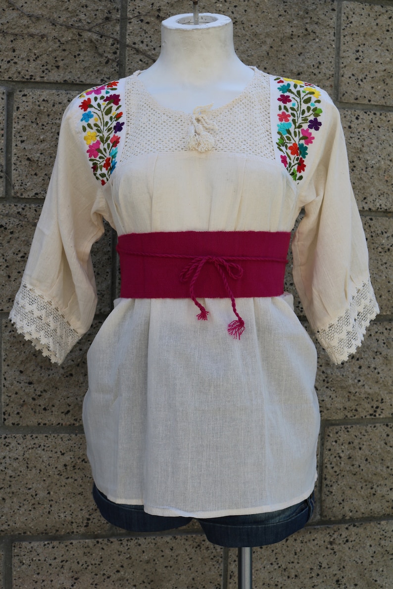 May include: A white cotton blouse with colourful floral embroidery on the sleeves and neckline. The blouse has a crochet neckline and a pink fabric belt.