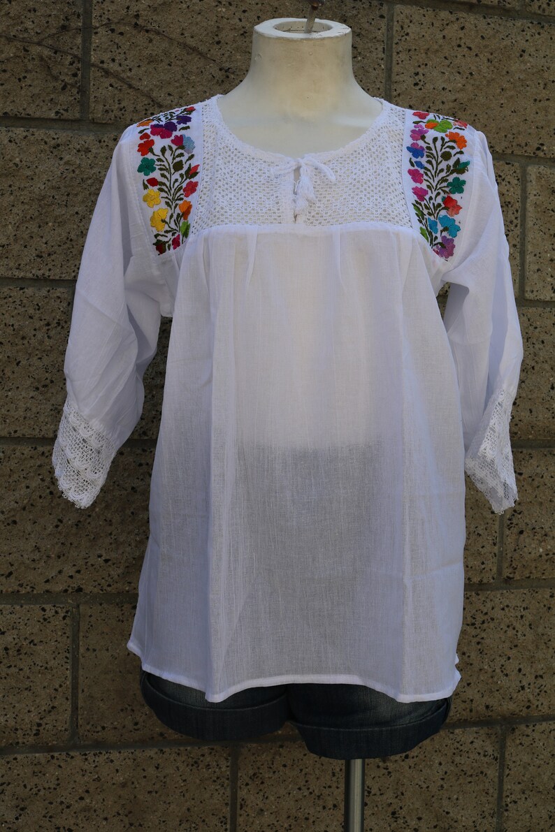 May include: White cotton blouse with colourful floral embroidery on the shoulders and a crocheted neckline. The blouse has 3/4 sleeves with crocheted trim.