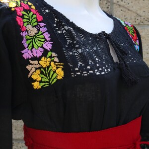 May include: Black embroidered blouse with a red woven belt. The blouse has a lace neckline and colourful floral embroidery.