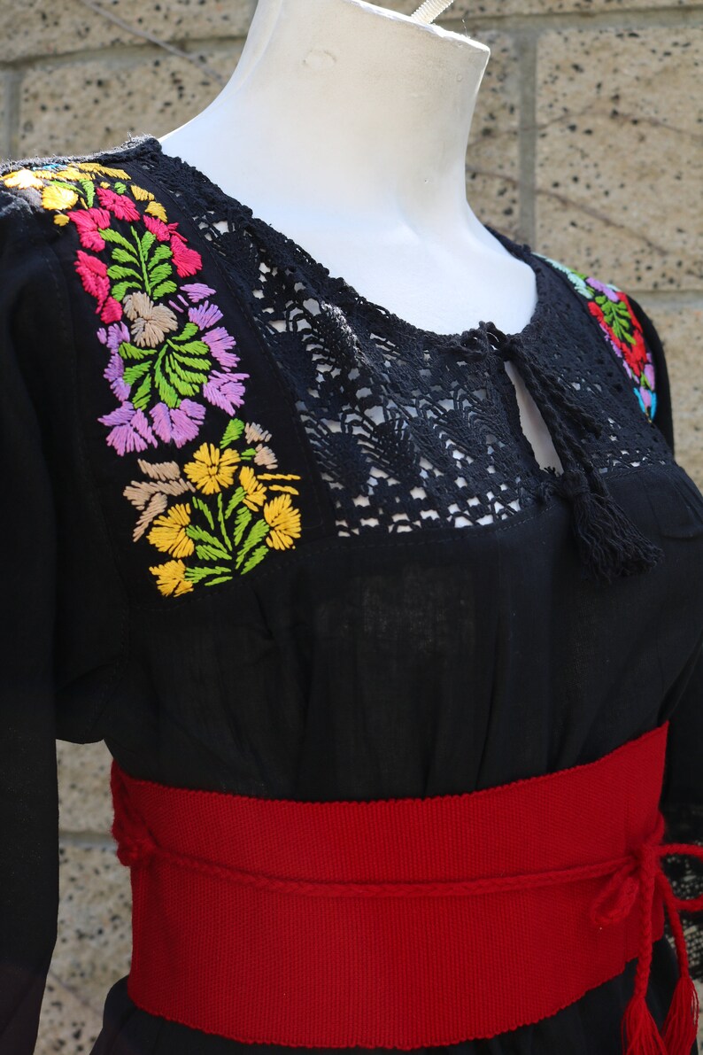 May include: Black embroidered blouse with a red woven belt. The blouse has a lace neckline and colourful floral embroidery.