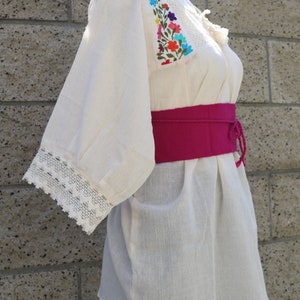 May include: A white cotton blouse with colourful floral embroidery on the neckline and sleeves. The blouse has a lace trim on the sleeves and a pink fabric belt.