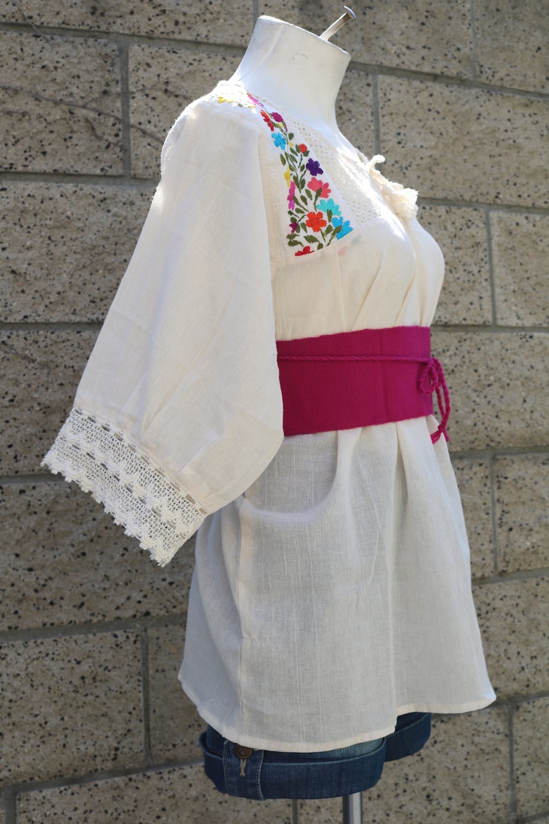 May include: A white cotton blouse with colourful floral embroidery on the neckline and sleeves. The blouse has a lace trim on the sleeves and a pink fabric belt.