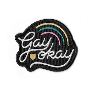 May include: Black embroidered patch with the words "Gay okay" in white script, a gold heart, and a rainbow design. The patch has a black border and is likely for clothing or accessories.