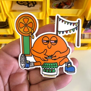 May include: A vibrant sticker depicting an orange character with closed eyes, seated and holding a small object. The sticker includes a lollipop, a banner with colourful flags, and a small cake. The sticker is held in a hand.