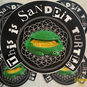 Puede incluir: Una pegatina en blanco y negro con el texto "This is Sande it Turtle" que rodea un recipiente verde con forma de tortuga lleno de material amarillo. La pegatina tiene un borde blanco y un patrón decorativo.