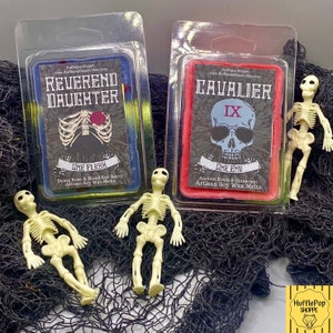 May include: Two wax melts in clear plastic packaging. The wax melts are red and blue. The red wax melt has a skull with sunglasses and the text "Cavalier IX One End Ancient Resin & Cinnamon Artisan Soy Wax Melts". The blue wax melt has a ribcage with a red heart and the text "Reverend Daughter One Flebh Dying Rose & Blood Red Berry Artisan Soy Wax Melts". Two plastic skeleton toys are in front of the wax melts.