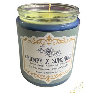 May include: A blue glass jar candle with a label that reads "GRUMPY X SUNSHINE". The label includes text about the scent, "Sweet Mango | Ozone | A Lil' Spice", and the candle's size, 227g. The candle has a yellow wax and a wick.