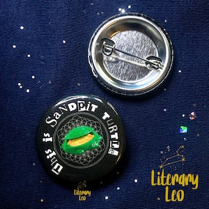 May include: Black and green button with the text "This is Sandpit Turtle" in white lettering. The button features a green turtle with a black and white mandala design in the background.