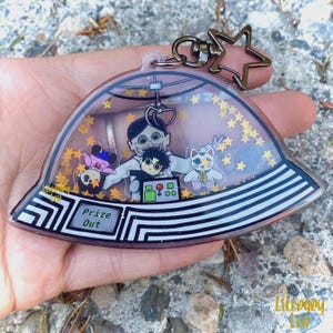 May include: A clear, dome-shaped keychain with a claw machine theme. Inside, cartoon characters and gold star-shaped glitter float. The keychain has a silver-colored metal star charm and the words "Prize Out" are visible.