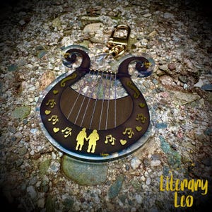 May include: A clear acrylic keychain in the shape of a lyre, featuring a brown center with gold musical notes, hearts, and silhouettes of two people holding hands. The keychain has a gold-colored clasp and the text "Literary Leo".