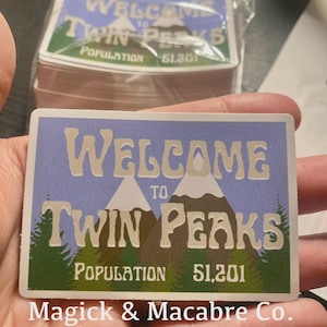 Könnte beinhalten: Ein rechteckiger Aufkleber mit blauem Hintergrund und den Worten "WELCOME TO TWIN PEAKS" in einer stilisierten Schriftart. Der Aufkleber zeigt Illustrationen von Bergen und Bäumen mit der Bevölkerungszahl 51.201. Der Text "Magick & Macabre Co." befindet sich unten.