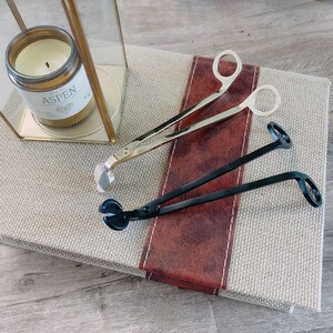 May include: Two candle wick trimmers, one gold and one black, on a beige surface with a red and brown stripe. The gold trimmer has a long handle and a curved end. The black trimmer has a long handle and a curved end with a loop.