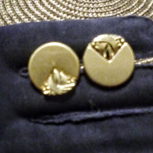 Puede incluir: Dos gemelos de color dorado con un diseño decorativo. Los gemelos son redondos y tienen un diseño en relieve en la parte delantera.