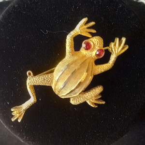 May include: A gold-toned frog brooch with red rhinestone eyes. The frog is in a leaping pose with its legs outstretched.