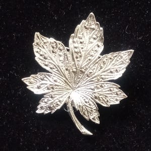 May include: A silver-toned leaf-shaped brooch with intricate detailing. The brooch features a detailed leaf design with small, sparkling accents. The brooch is set against a dark background, highlighting its metallic sheen.