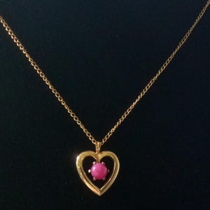 May include: A gold-colored necklace with a heart-shaped pendant. The pendant features a pink gemstone set within the heart. The chain is delicate and the background is black.