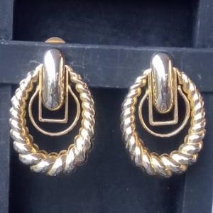 May include: A pair of gold-coloured earrings. Each earring has a twisted rope design around a central geometric shape and a rectangular top. The earrings are designed for pierced ears.