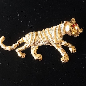 May include: A gold-toned tiger brooch with red rhinestone eyes and a clear rhinestone body. The brooch is set on a black background.