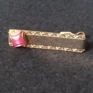 May include: A gold-coloured tie clip with a rectangular form and a decorative edge. The clip has a dark centre and a pink, geometric accent on one end. The tie clip is designed to secure a tie to a shirt.