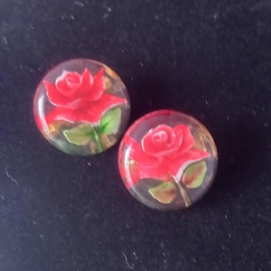 May include: Two round, clear earrings with a red rose design. The roses have green leaves and are set against a gold and brown background. The earrings are on a dark background.