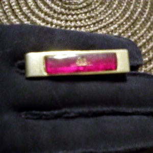 May include: A gold-toned bar with a red glass insert. The glass insert has a small gold design in the center.