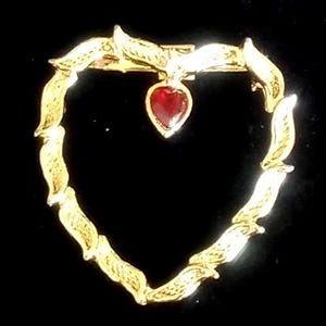 May include: A gold-toned heart-shaped brooch with a red heart-shaped gemstone pendant. The brooch is made of a series of stylized leaf shapes. The background is black.