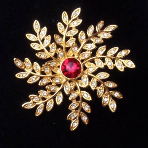 May include: A gold-toned snowflake brooch with a central, round, red gemstone. The brooch features delicate leaf-like designs, each adorned with small, clear crystals. The background is black.