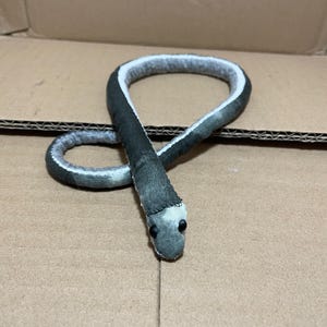 May include: A plush snake toy crafted from dark grey fabric with a lighter grey underside and a white stripe running along the length of its body. The snake features two small black bead eyes and is coiled into a loop on a flat surface.