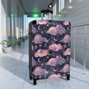 May include: A rolling suitcase with a whimsical night sky design. The navy blue background is adorned with pink and purple clouds, gold crescent moons, and stars. The suitcase has a black frame and handle.
