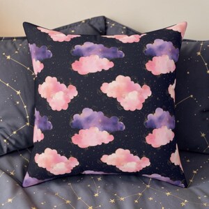 May include: Square decorative pillow with a dark blue background and a pattern of pink and purple clouds. The clouds have gold accents and are scattered across the pillow. The pillow is on a bed with a constellation pattern.