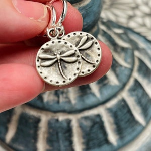 May include: Silver hoop earrings with a dragonfly charm. The dragonfly is detailed and has a textured finish.