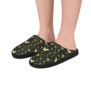 Peut inclure: Paire de pantoufles vert foncé avec un motif répété de papillons, de champignons et de croissants de lune dorés et crème. Les pantoufles ont une semelle noire et sont conçues pour un usage intérieur, offrant confort et style.