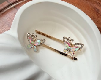 May include: Two gold-toned bobby pins with iridescent butterfly embellishments. The butterfly designs are adorned with small, clear rhinestones. The bobby pins are displayed on a white, seashell-shaped dish.