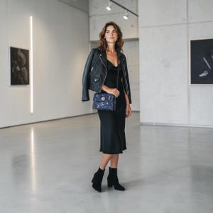 May include: A woman in a black dress and leather jacket stands in a gallery. She carries a small, dark blue crossbody bag with a crescent moon and star pattern. She wears black ankle boots.