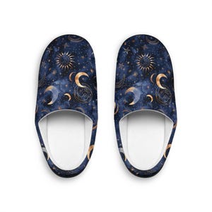 May include: A pair of celestial-themed slippers. The slippers feature a dark blue background with a pattern of gold moons, stars, and suns. The interior is white. These cozy slippers are designed for indoor use.