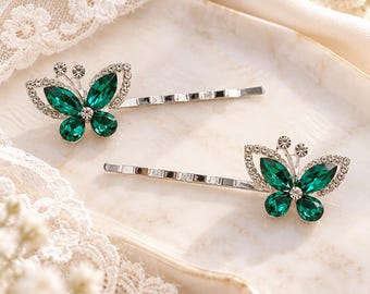 May include: Two silver-toned hair clips, each featuring a butterfly-shaped ornament. The butterfly wings are crafted from deep emerald green faceted crystals, bordered by small clear rhinestones. The metal hair pins have a simple, straight design.