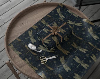 May include: A wrapped gift with a brown ribbon sits on a round wooden table, surrounded by matching wrapping paper. The paper features a dark blue background with a gold dragonfly and constellation pattern. Scissors and tape are also on the table.