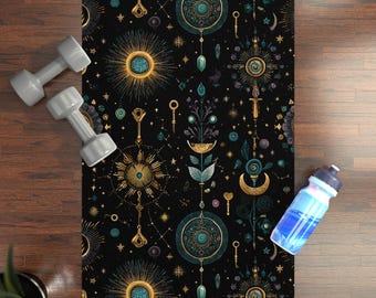 Op de afbeelding: Een zwarte yogamat met een hemels ontwerp met zonnestralen, sleutels en geometrische vormen in goud en blauwgroen. Twee grijze dumbbells en een blauwe waterfles liggen naast de mat. De mat ligt op een houten vloer.