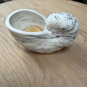 May include: A seashell-shaped candle holder made of white and gray marbled material. The candle holder has a small, round opening to hold a candle. The candle is a light yellow color. The holder sits on a light brown wooden surface.
