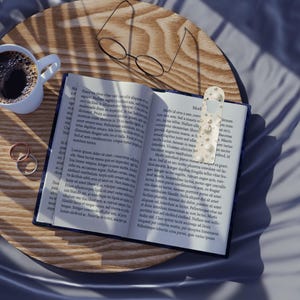 May include: A close-up of a book with a bookmark, reading glasses, a coffee cup, and two rings on a wooden tray. The book is open, and the bookmark has a white background with small, decorative shapes. The scene is lit by soft light.
